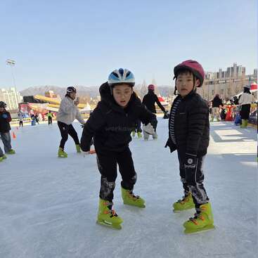Deux enfants portant des casques et des patins à glace verts font du patin à glace en plein air. D'autres personnes patinent à l'arrière-plan, avec des bâtiments et des montagnes sous un ciel bleu clair.