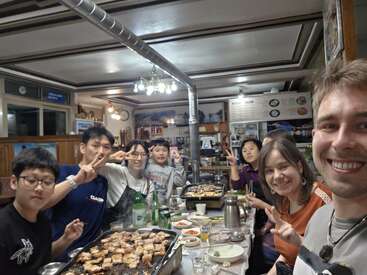 Eine Gruppe von Leuten genießt gemeinsam ein koreanisches Barbecue in einem Restaurant, lächelt und macht Peace-Zeichen, umgeben von leckerem Essen, Getränken und einer herzlichen Atmosphäre.