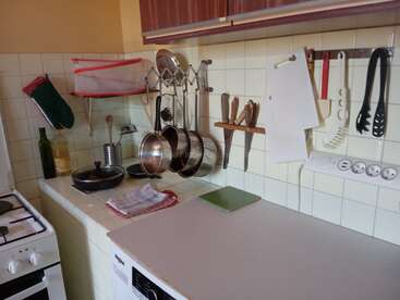 Esta imagen muestra un rincón de cocina ordenado con ollas colgantes, utensilios, tablas de cortar, cuchillos, manopla de horno, aceite de oliva y diversos utensilios de cocina organizados contra las paredes de azulejos.