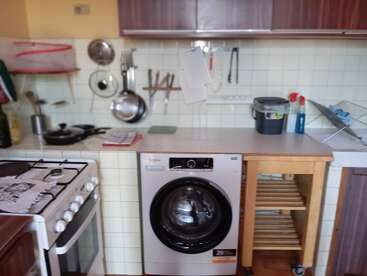 Esta imagen muestra una cocina compacta con lavadora, fogones, ollas y utensilios colgados, encimera con artículos de limpieza, carrito de madera, tendedero y salpicadero de azulejos.