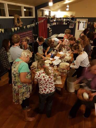 Un groupe de personnes est réuni autour d'une grande table en bois ; dans une cuisine chaleureuse et bien éclairée, elles participent avec enthousiasme à un cours de cuisine et échangent des recettes.