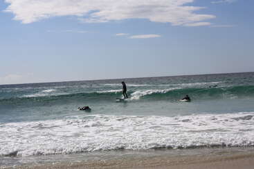 Três surfistas aproveitam as ondas do mar sob um céu parcialmente nublado. Um deles está surfando uma onda, enquanto os outros esperam ou remam nas águas lindas e cristalinas.