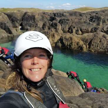Uma mulher sorridente, usando capacete e roupa de mergulho, tira uma selfie à beira de águas cristalinas de cor turquesa, com falésias rochosas e outras pessoas em roupas de mergulho ao fundo.