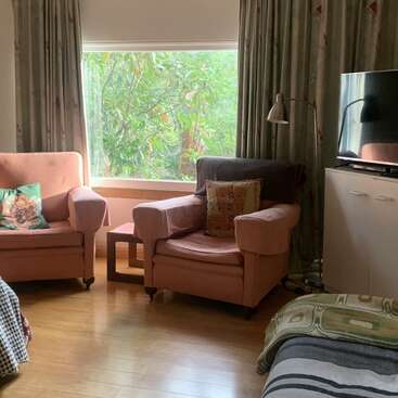 Cette chambre confortable dispose de deux fauteuils roses avec des coussins colorés, d'une grande fenêtre avec vue sur la verdure, de parquet, d'une télévision et d'un éclairage naturel doux. Atmosphère calme.