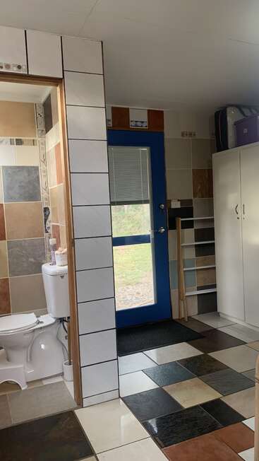 Un cuarto de baño con azulejos de colores y una entrada con una puerta azul, un inodoro blanco, un armario y varios enseres domésticos. La luz natural entra a raudales desde el exterior.