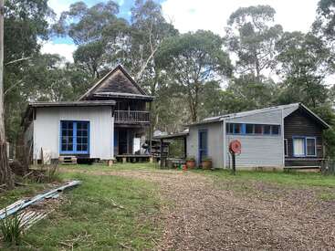 Dos casas rústicas con revestimiento metálico y ventanas con ribetes azules se alzan entre altos árboles. El patio es de hierba, con un camino de grava, macetas esparcidas y muebles de exterior.