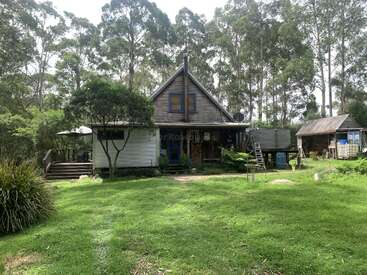 Una casa rústica de madera rodeada de altos árboles y exuberante vegetación, con una terraza, muebles de exterior y un pequeño cobertizo en un entorno rural tranquilo.