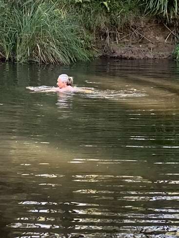 Una persona con el pelo canoso nada en un estanque sereno y natural rodeado de hierba alta y verde y orillas terrosas, disfrutando de un momento de paz y tranquilidad al aire libre.