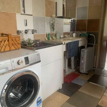 Esta imagen muestra un lavadero alicatado con una lavadora, un fregadero, un calentador, una cesta, artículos de limpieza y un espejo. Toallas y flores decoran el espacio.