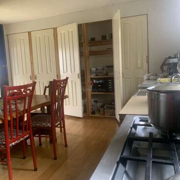 Una acogedora cocina con suelos de madera, sillas rojas, mesa de madera, puertas de despensa abiertas que dejan ver estanterías con utensilios de cocina, un fogón y varios elementos de encimera crean una escena hogareña.