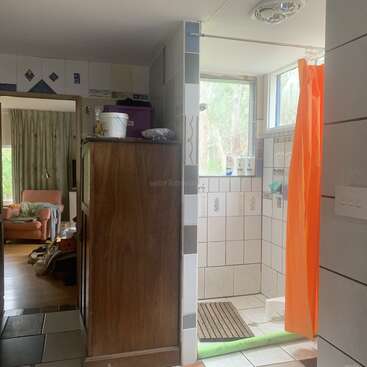 La acogedora vivienda cuenta con un cuarto de baño alicatado con una cortina de ducha de color naranja chillón, un armario de madera y una sala de estar contigua con un sillón y enseres dispersos.