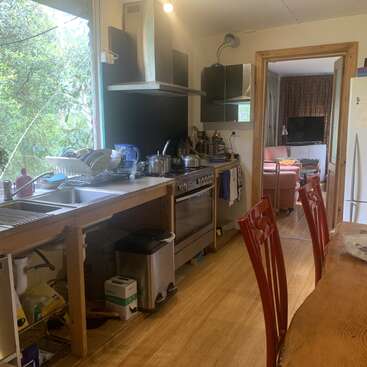 Una acogedora cocina con mesa de madera, sillas rojas, electrodomésticos de acero inoxidable y una gran ventana. Se abre a un salón con TV y sillón.