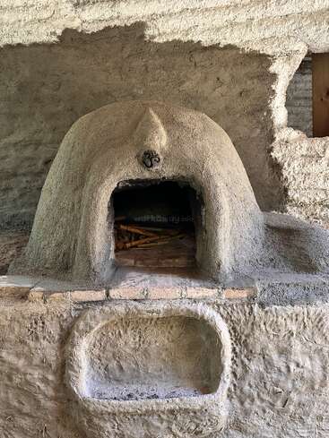 This image shows a rustic, dome-shaped clay oven with an arched opening, built into a textured cob wall, featuring an Om symbol and a built-in shelf below.