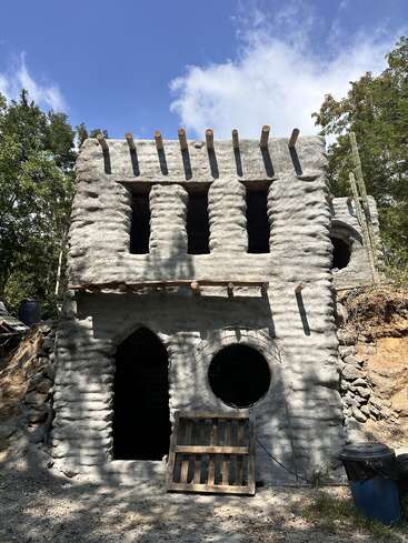 The image shows a rustic, unfinished two-story building made of rough cement with arched and circular windows, wooden roof beams, surrounded by trees and blue sky.