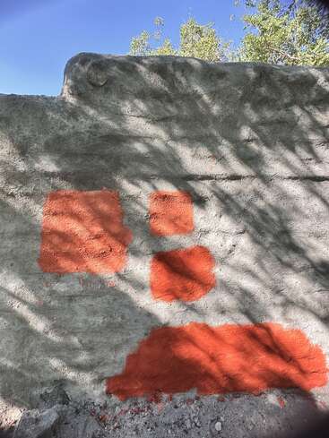 A rough concrete wall features four red-orange paint patches of varying sizes. Shadows from nearby trees fall across the surface, with a clear blue sky above.