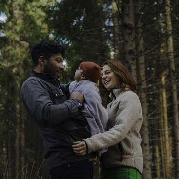 The image depicts a family of three, consisting of a mother, father, and child, posing for a photo, with the mother and father standing on either side of their child.