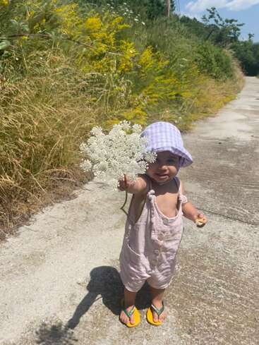 Un niño pequeño, con un sombrero para el sol y sandalias, está de pie en un sendero bañado por el sol, sosteniendo un ramo de flores silvestres blancas, rodeado de hierba alta.