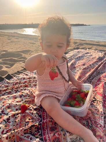 Un niño pequeño está sentado en una manta estampada en la playa, con una fresa en la mano. El sol se pone sobre el mar, proyectando un cálido resplandor dorado.