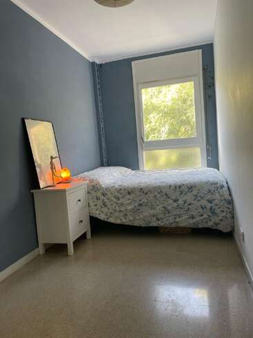 Una habitación pequeña y acogedora con paredes azules y blancas, una cama individual con ropa de cama a flores, una ventana, una mesita de noche, un cuadro enmarcado y una lámpara encendida. Ambiente tranquilo.