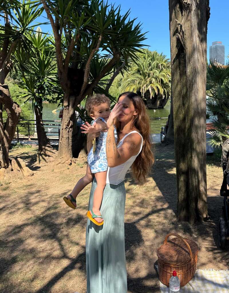 Una mujer con una camiseta blanca y una falda larga sostiene a un niño en un parque. Se ven árboles, una cesta de picnic y una botella de agua, con la luz del sol y la vegetación de fondo.