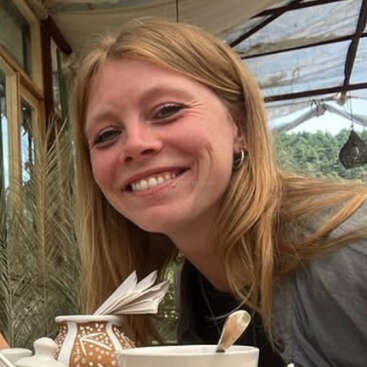 L'image représente une femme aux longs cheveux blonds souriant à l'appareil photo, assise à une table avec une tasse à thé blanche et un vase décoratif brun et blanc.