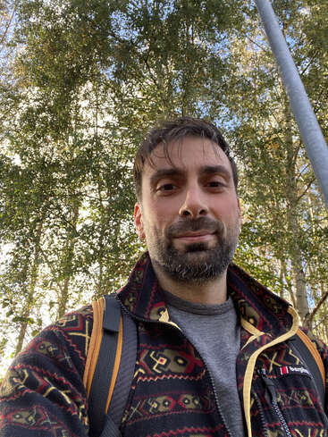 Ein bärtiger Mann macht draußen ein Selfie; er trägt eine gemusterte Jacke und einen Rucksack. Sonnenlicht fällt durch die hohen grünen Bäume hinter ihm und schafft eine angenehme Atmosphäre.