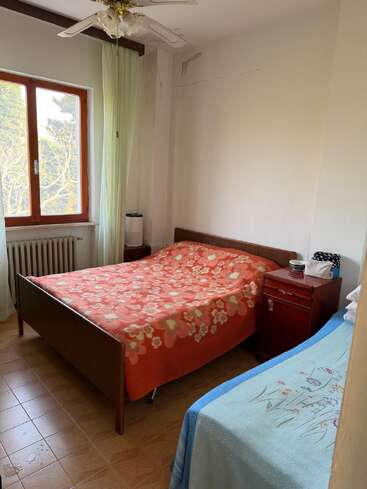 Dieses gemütliche Schlafzimmer verfügt über ein Doppelbett mit einer orangefarbenen Decke mit Blumenmuster, ein Einzelbett mit einem blauen Bezug, Holzmöbel, einen Deckenventilator und ein Fenster.