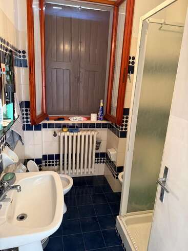 Dieses Bild zeigt ein kompaktes Badezimmer mit blau-weißer Fliesenverkleidung, einer Duschkabine, einem Waschbecken, einer Toilette, einem Bidet, einem Heizkörper und einem Fenster mit Holzläden.
