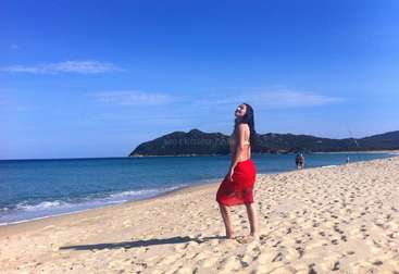 A woman stands on a beach, wearing a dress, with a body of water behind her. The background is a sandy beach with a clear blue sky. The overall atmosphere is one of relaxation and tranquility.