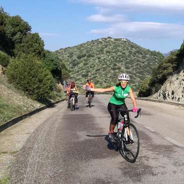 The image depicts a group of cyclists riding on a scenic road, surrounded by trees and hills, with one cyclist extending her arm in a friendly gesture.