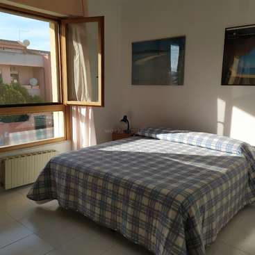 The image depicts a serene bedroom with a double bed, white tiled floor, and a large window offering a view of the outside. The room is well-lit.