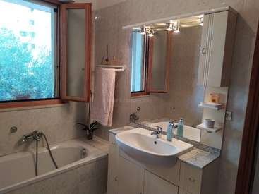 The image depicts a bathroom with a bathtub, sink, and mirror, featuring white cabinets, a window, and a towel rack, with a plant on the counter.