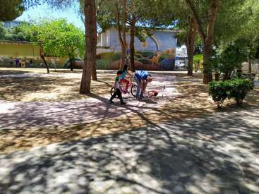 The image depicts two children in a park, one standing and the other bent over a bicycle, surrounded by trees and a mural on a building in the background.