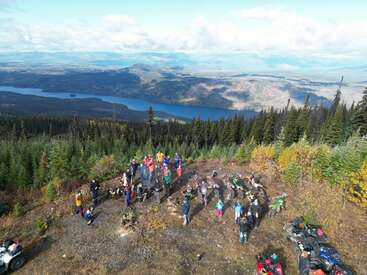 Um grupo de pessoas se junta num mirante na montanha, cercado por florestas e folhagem colorida de outono, com motos e quadriciclos, com vista para um grande lago panorâmico.