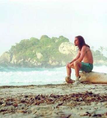 A man with long hair sits shirtless on a log at the beach, gazing thoughtfully at the ocean with an island covered in greenery behind him.