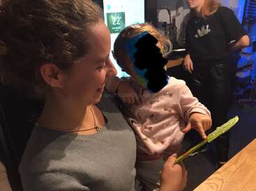 A young woman smiles while holding a small child. The child’s face is obscured. Another woman stands in the background. The setting appears to be a kitchen.