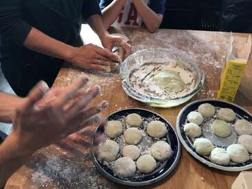 Three people are making mochi by hand at a wooden table, shaping soft dough into round balls, dusting them with flour, and placing them on trays.
