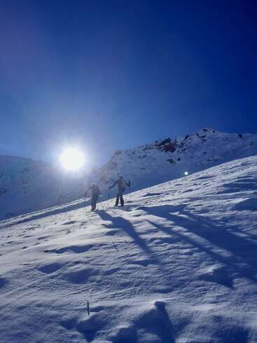 Two people hike uphill through thick snow on a sunny, clear day. Long shadows stretch behind them, surrounded by snowy mountains and vibrant blue sky.