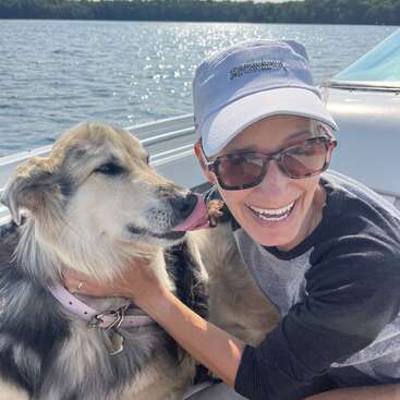 Uma mulher alegre, usando óculos escuros e boné, está sorrindo em um barco, abraçando um cachorro feliz com uma coleira rosa. Eles estão curtindo um dia ensolarado juntos.
