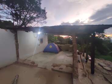 Una tienda azul se instala en el interior de los restos de una antigua estructura con paredes y techo parciales, rodeada de naturaleza y árboles al atardecer.