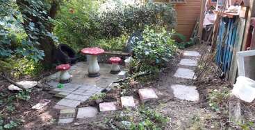 Esta imagem mostra uma área de jardim extravagante com mesa e banquetas em forma de cogumelo, cercada por vegetação, pedras de pavimentação e um galpão de madeira, criando um ambiente aconchegante na floresta.
