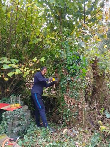A person wearing dark clothes and gloves is sawing a tree trunk in a lush, overgrown garden. Tools and green foliage surround them, indicating outdoor work.