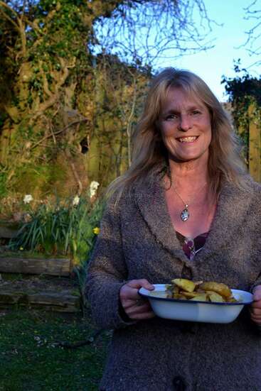 The image depicts a woman holding a white bowl of food in a garden setting, with long blonde hair and a brown cardigan, smiling at the camera.