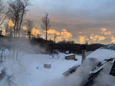 Uma paisagem de inverno com neve ao pôr do sol, com nuvens alaranjadas no céu, vapor subindo dos telhados, árvores nuas e um pequeno galpão à distância.