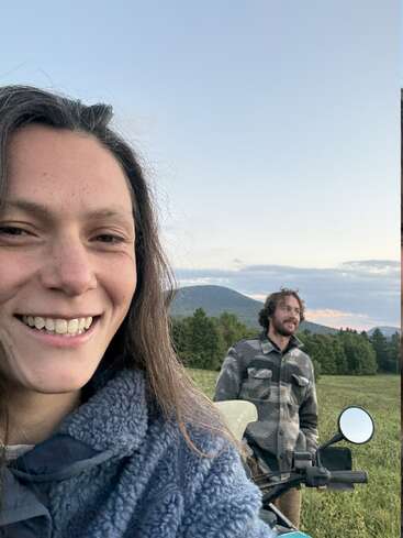 Uma mulher sorridente tira uma selfie ao ar livre, com um homem atrás dela ao lado de uma motocicleta. Eles estão em um campo verde com árvores e montanhas.