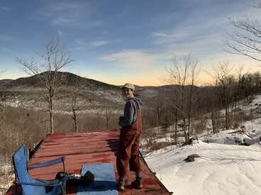 Uma pessoa está em uma plataforma vermelha com vista para montanhas nevadas e árvores nuas ao pôr do sol. Uma cadeira e uma mochila azuis descansam nas proximidades, criando uma visão pacífica e cênica.