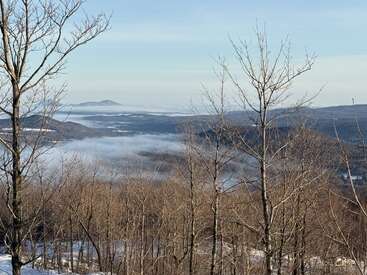 Árvores sem folhas aparecem em primeiro plano. Colinas onduladas e vales enevoados se estendem à distância. Uma paisagem serena de inverno sob um céu azul claro, pacífico e calmo.