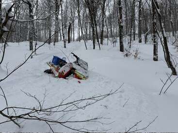 Um snowmobile parece estar preso ou abandonado na neve profunda em uma floresta de inverno tranquila e sem folhas, cercado por árvores nuas e neve branca intocada.