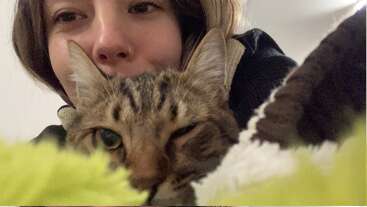 Uma selfie em close-up mostra uma pessoa sorridente com um piercing no nariz abraçando um gato malhado relaxado. Ambos parecem confortáveis e satisfeitos, deitados juntos em cobertores macios.