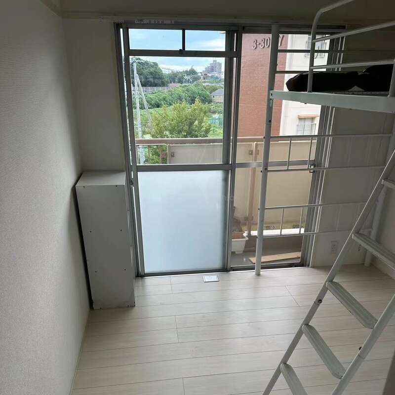 Das ist ein kleines, minimalistisch eingerichtetes Zimmer mit hellem Holzboden, einem Hochbett mit Leiter, einem großen Fenster, einem Balkon und Blick auf die Stadt und die grüne Umgebung.